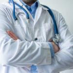 Photo of body of doctor in white lab coat with stethoscope on his neck in half-turn with folded hands against white wall in hospital with light source on left. Concept image of  medical worker